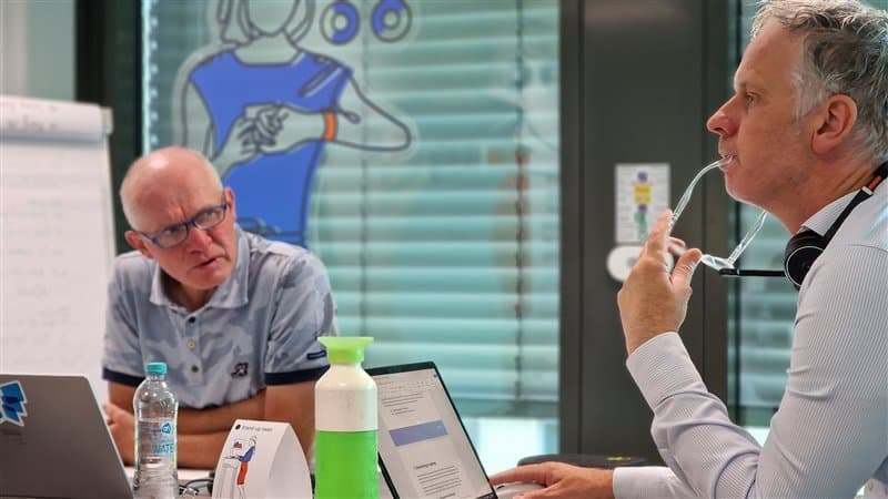 Two men at a table with laptops discussing in a modern office with a mural in the background.